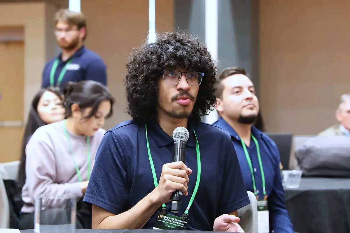 A student in a navy polo holds a microphone and asks a question during a talk, with other attendees seated behind him.
