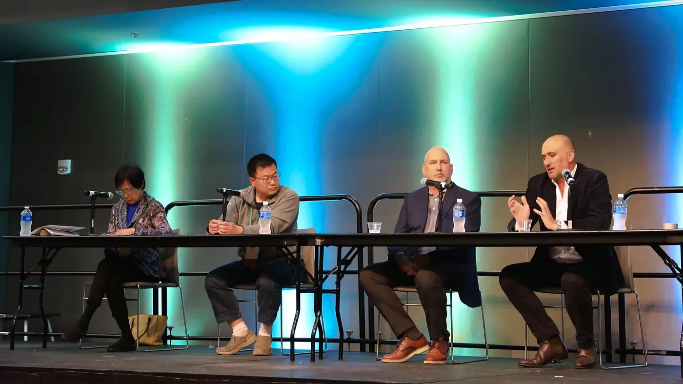 Four panelists sit at a table on stage with microphones while one gestures as he speaks, against a blue and green lit backdrop.