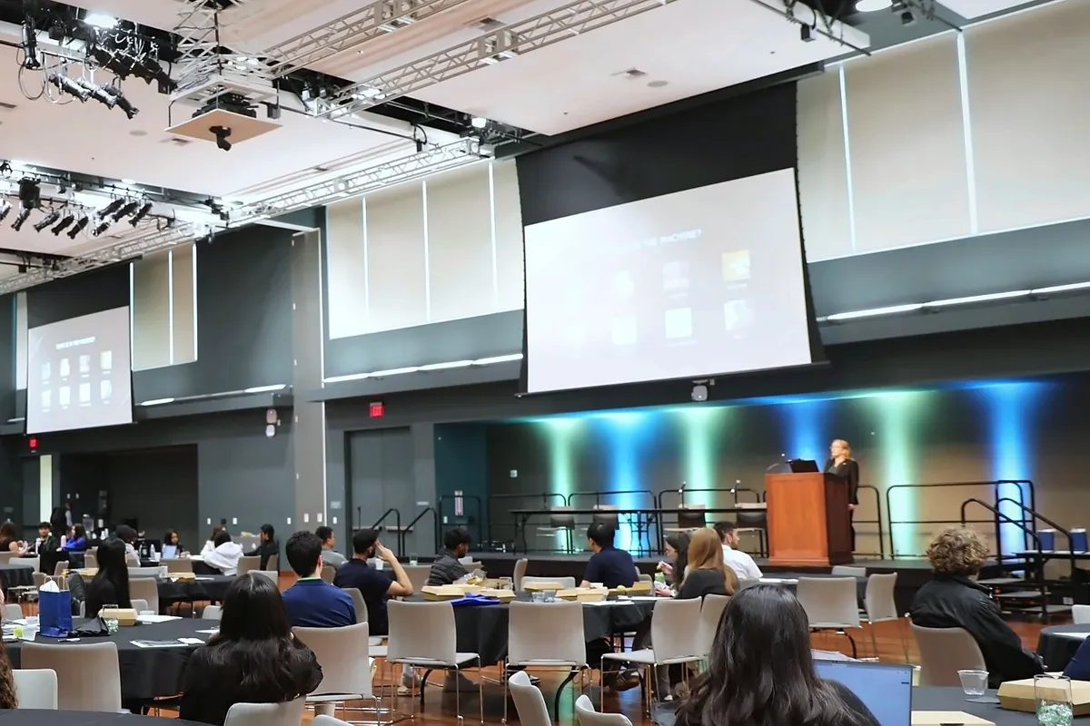 Keynote speaker Kate Stephenson, PhD, presents “Accounting for AI in Future BME Careers” at a podium on stage beside two large screens while attendees sit at round tables.