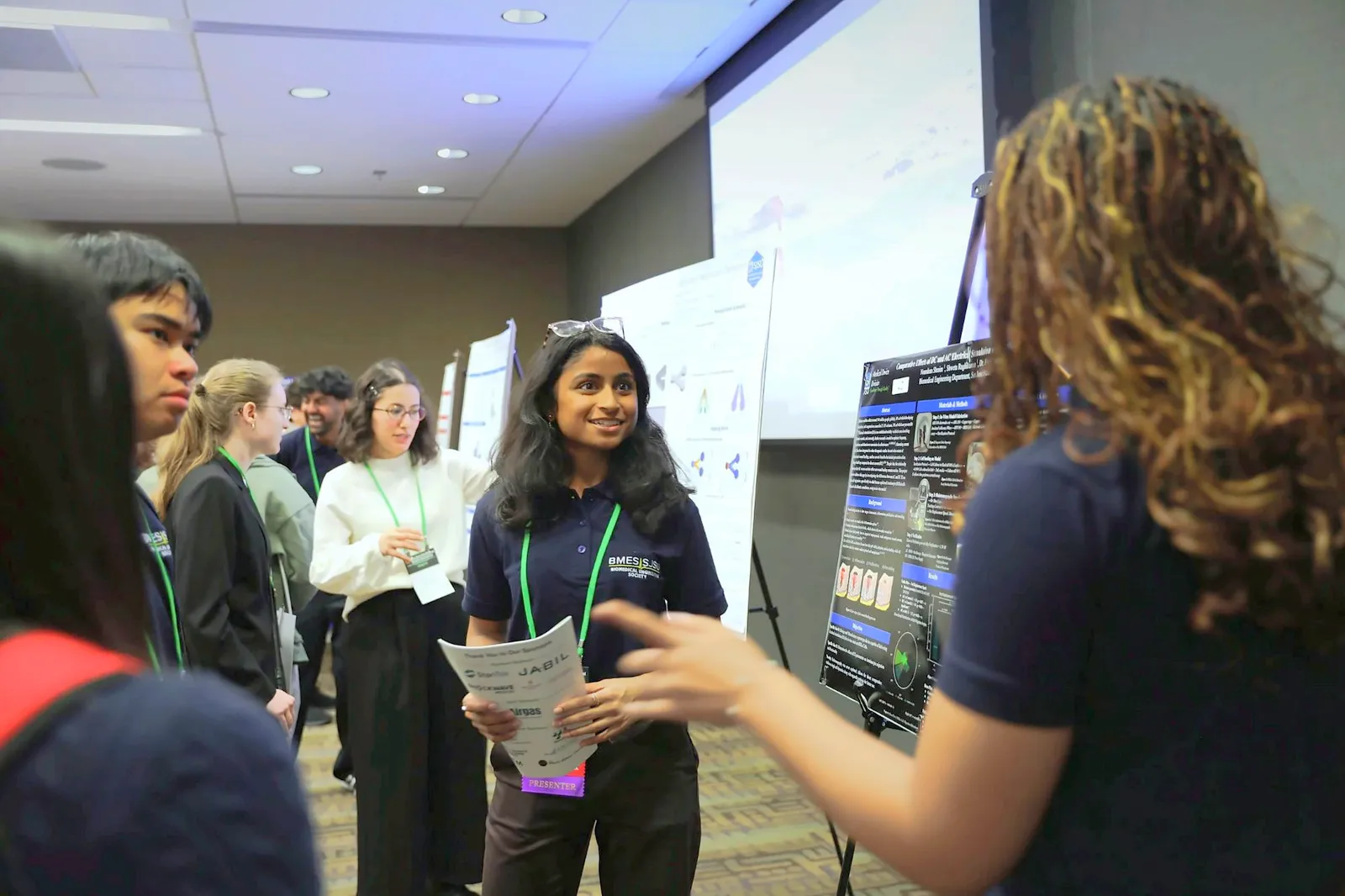 A presenter wearing a lanyard and holding a program talks with attendees in front of research posters.