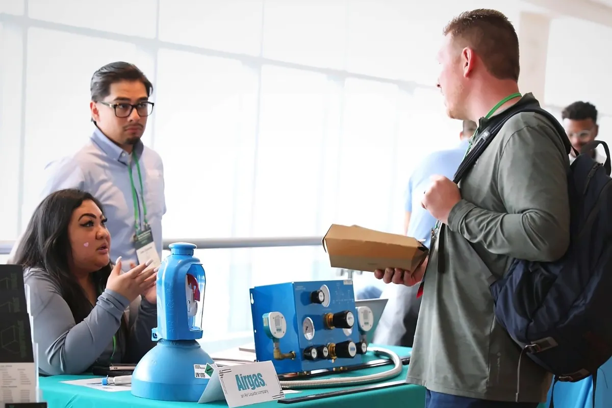 Two Airgas representatives at a table with medical gas equipment talk with an attendee wearing a backpack.